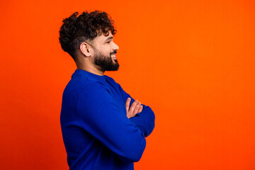 Confident smiling man in blue sweater standing in profile with arms crossed against bright orange background © deagreez