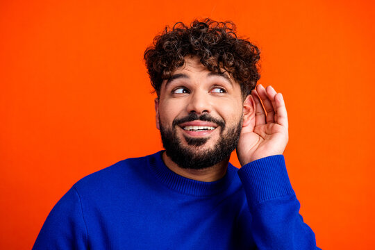Young man listening intently with hand to ear in bright portrait, curious expression, blue sweater against orange background conveying communication and attentive listening