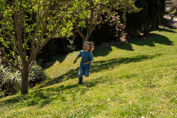 Fototapeta premium Little blond boy running on green hill in sunny park, childhood freedom concept, outdoor activity, joyful movement, spring nature, lifestyle scene, carefree play, landscape background