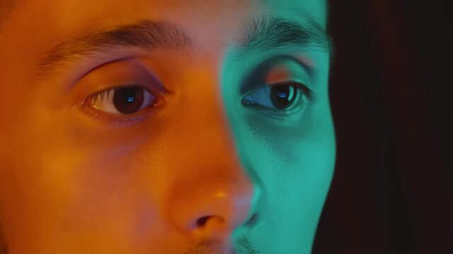 A man's face is bathed in split orange and teal lighting as he focuses intently, his eyes darting and blinking while staring at a screen in the darkness.