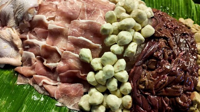 Close-up of fresh raw pork slices, chicken, pork liver and meatballs neatly arranged on a banana leaf at a Thai street food market. Traditional ingredients prepared for barbecue, hot pot and local cui