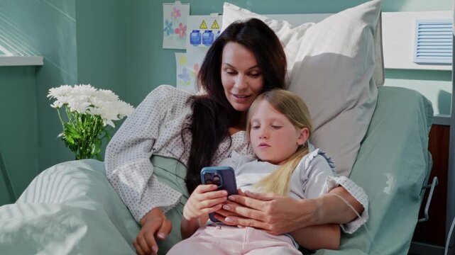 Mother and daughter use smartphone lying together on hospital bed