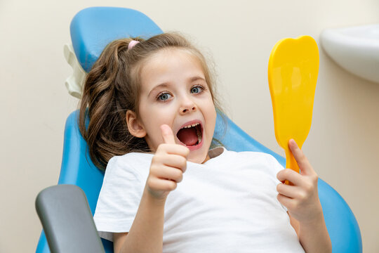 Portrait of a cute happy little girl sitting in dental chair smiles and gives thumbs up while holding a yellow dental mirror in a bright clinic environment