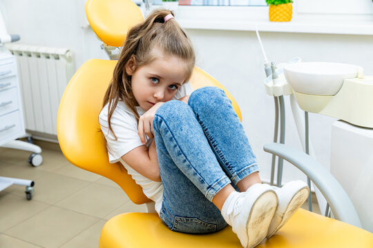 Childish fear. Child girl with brown hair sits curled up in a yellow dental chair, appearing apprehensive in a well-lit dental office featuring modern equipment, a window in the background