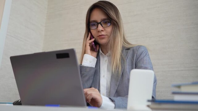 the man is talking on two phones at once. Businessman multitasking on a phone call while reaching for papers and a pen at his office desk. High workload and busy day.
