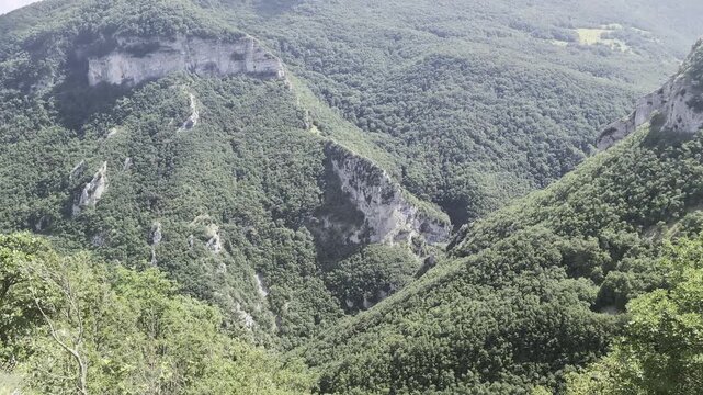 Paesaggio montano con pendii ricoperti di vegetazione fitta, rocce affioranti e vallate profonde che si estendono verso l&rsquo;orizzonte, sotto un cielo parzialmente nuvoloso