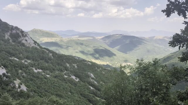 Paesaggio montano con colline verdi ricoperte di vegetazione fitta, alberi e arbusti in primo piano e una serie di vallate che sfumano verso l&rsquo;orizzonte sotto un cielo parzialmente nuvoloso