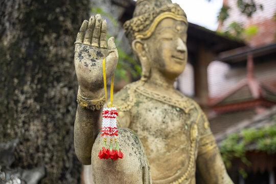 Stone guardian statue with raised hand holding red and white floral garland, serene temple scene conveying peaceful devotion and cultural tradition