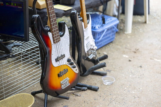 Sunburst electric bass guitar and black electric guitar on stands at outdoor market with music gear and crate, casual scene with warm tones and relaxed atmosphere
