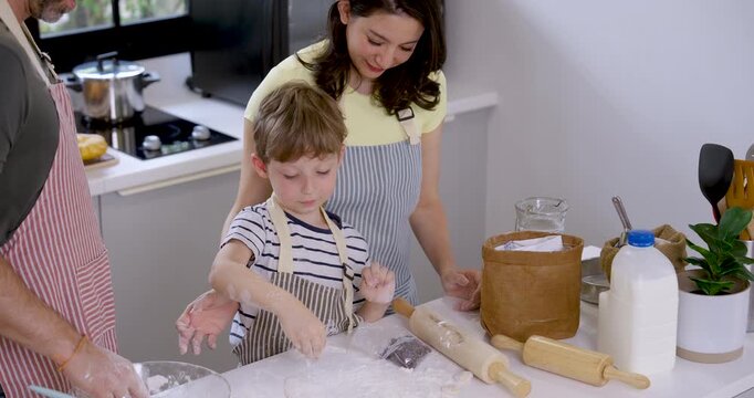 Caucasian young son reaches into flour container and spreads flour across kitchen counter while parents guide preparation, teaching home baking skills through patient parenting and family teamwork.