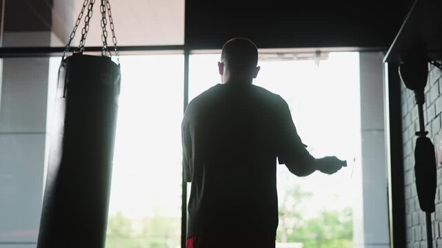 Backlit silhouette of athlete holding jump rope inside gym, framed by punching bag, window light, brick wall, strong contrast, training atmosphere, focused stance, sporty interior
