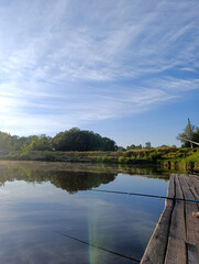 Fototapeta premium Fishing rods on a wooden pier by a calm river on a sunny morning