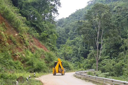 Yellow excavator backhoe driving on winding mountain road surrounded by dense tropical forest and green hillside, peaceful rural construction scene with guardrail and overcast sky