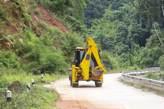 Yellow excavator backhoe parked on winding mountain road surrounded by green forest and steep hillside, construction vehicle waiting beside guardrail under overcast sky