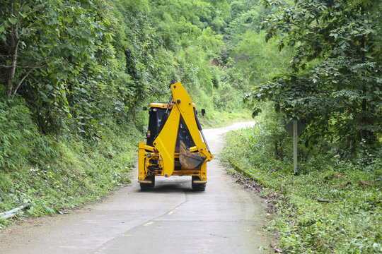 Yellow excavator parked on narrow forest road surrounded by dense green foliage, conveying quiet work readiness and rural construction scene