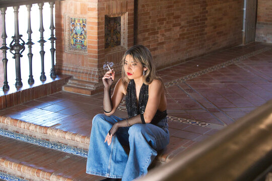 A young, attractive woman wearing jeans, gloves and a black T-shirt, smoking a cigarette whilst sitting on the steps of Spain Square in Seville. The woman is on holiday in the city.