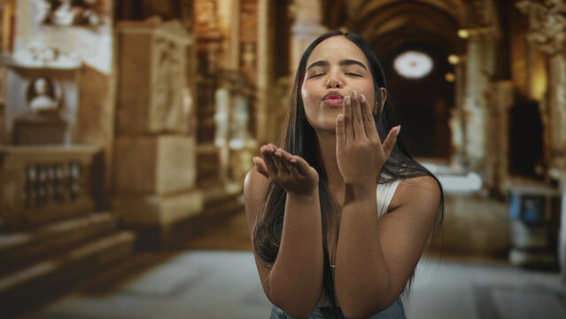 Young hispanic woman blows a kiss with raised hand to lips in an ornate building hall, puckered lips and casual denim visible; affection warmth.