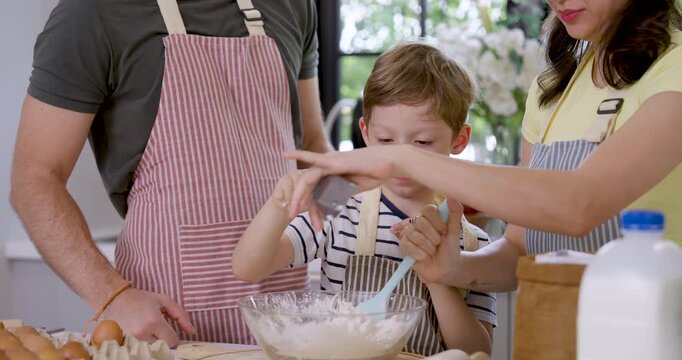 Caucasian mother pours liquid ingredient into glass bowl while young son whisks flour mixture and father stands nearby, creating home baking lesson with family teamwork, parenting and kitchen bonding.