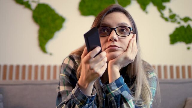 Bored woman holding a remote control, switching TV channels with a dull expression. Tedious television, apathetic viewing, and lack of interest.