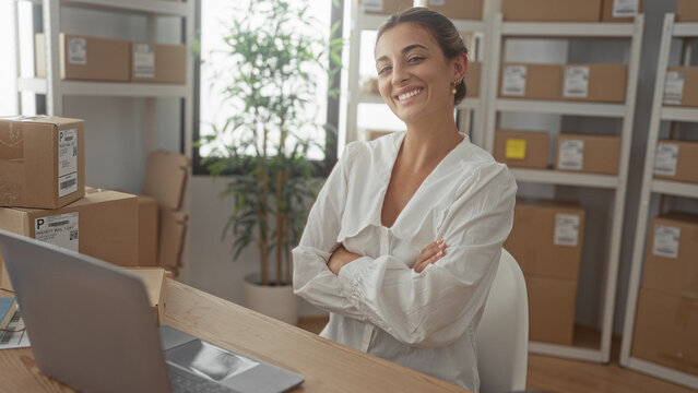 Woman smiling and typing on laptop among stacked parcels and shipping boxes in a light filled building; small business confidence.