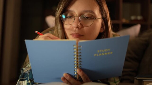 Smart student young woman writing in a notebook while lying on a sofa with a stack of books. Home studying, creative writing, and cozy student lifestyle. writing a curriculum.