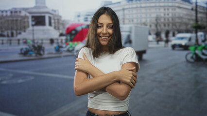 Fototapeta premium Young hispanic woman hugging bare shoulders with arms crossed on busy city street among passing vans and buses; self care contentment.