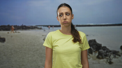 Fototapeta premium Woman pouting face at beach in yellow t shirt, ponytail and standing posture with shoreline rocks visible; boredom holiday travel.