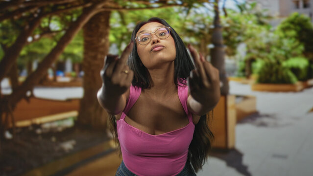 Young hispanic woman in pink tank top shows middle fingers toward the camera on a street plaza; defiance attitude.