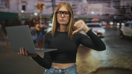 Fototapeta premium Woman holding laptop and pointing finger on a busy street in front of cars and buildings; frustration critique.