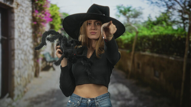 Woman in witch hat holds a large black spider prop with one hand, points her finger directly at the viewer while showing a bare midriff in a crop top and jeans on a street; mischief.