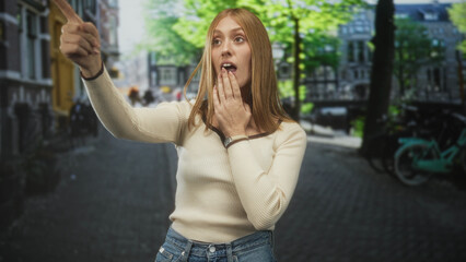 Fototapeta premium Woman pointing with outstretched arm and hand to mouth gesture by a bicycle on a cobblestone street, searching for direction; surprise discovery.
