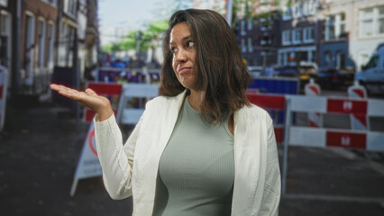 Fototapeta premium Woman shrugging with palm up hand at a blocked urban street with construction barriers and vans, looking unimpressed; frustration inconvenience.
