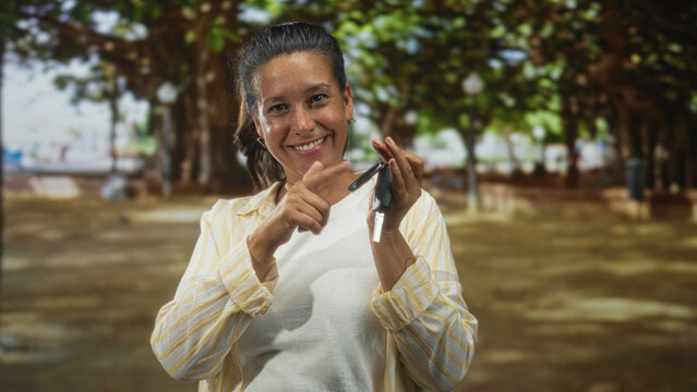 Hispanic middle age brunette woman holding car keys and key fob, smiling and makes call hand gesture on street; friendly invitation.