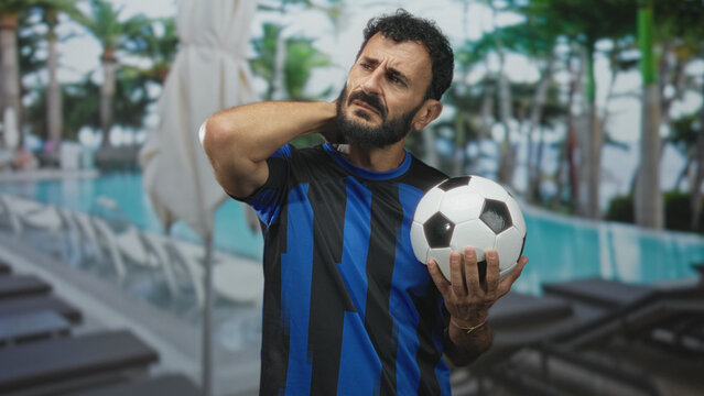 Man holding soccer ball with hand on neck and grimace at building poolside near lounge chairs and umbrella; injury pain recovery.