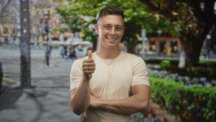 Fototapeta premium Young man with glasses giving thumbs up and arms crossed on a city street near trees and flower beds, smiling and clasping hands; friendly confidence.