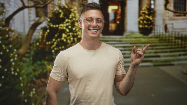 Man points finger to building entrance steps while smiling, wearing glasses and beige tshirt, visible forearm; cheerful welcome.