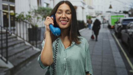 Fototapeta premium Woman holding a blue telephone handset to her ear on a city street, smiling, young brunette hispanic; urban connection happy.