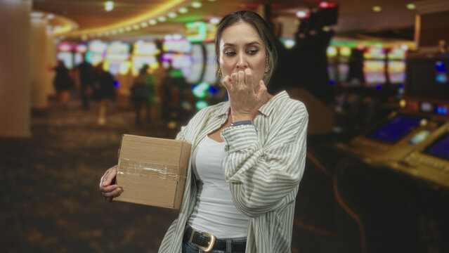 Woman holding a cardboard box while biting her fingernail and glancing sideways in a building; anxiety hesitation.