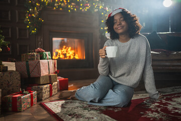 Obraz premium Merry Christmas. African American woman in Santa hat with cup of hot drink coffee tea near Christmas tree in dark interior. Girl in living room with Christmas tree and fireplace. Christmas eve at home