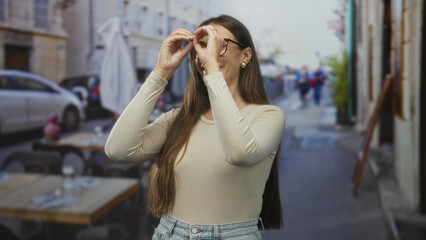Fototapeta premium Woman forms binoculars with hands over her eyes at an outdoor street cafe beside tables and parked cars, wearing glasses and denim; curiosity discovery.
