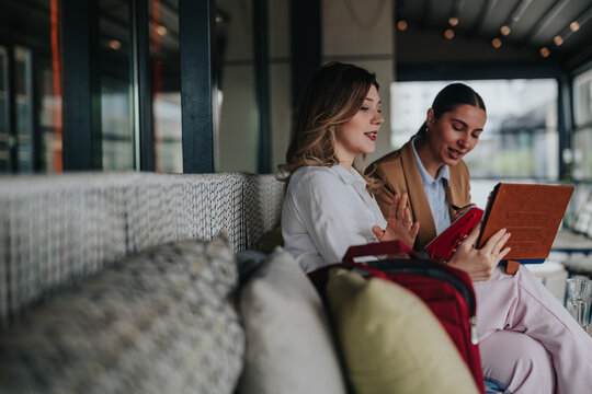 Two female colleagues review documents and discuss ideas using a tablet while working remotely. The pair collaborate in a relaxed cafe setting for a casual business meeting.