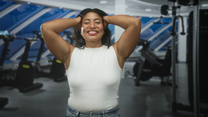 Fototapeta premium Woman in white sleeveless top with hands behind head and hair tossed, smiling in gym building among cardio machines; joy relief celebration.