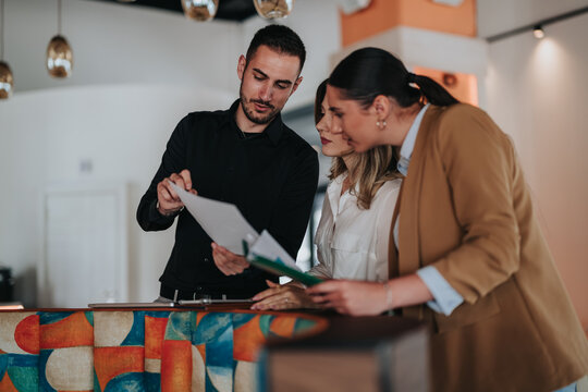 Three colleagues review paperwork together at a modern office reception desk. The team discusses documents while coordinating remote work and schedules.