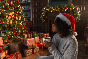 Obraz premium Merry Christmas. African American woman in Santa hat with cup of hot drink coffee tea near Christmas tree in dark interior. Girl in living room with Christmas tree and fireplace. Christmas eve at home