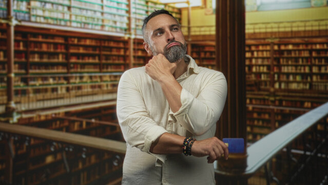 Middle aged hispanic man holding blue creditcard with hand on chin in building library surrounded by bookshelves; contemplation decision doubt.