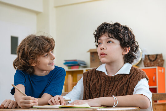 Two boys sit together in classroom, one boy politely asks a friend with smile while his classmate focuses ahead attentively, showing genuine communication and peer interaction in elementary school