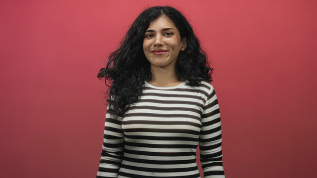 Young arab woman wearing a striped top smiles with eyes closed showing cheek piercings and bare face in a pink studio setting; contentment.