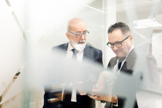 Shot through a glass of two businessmen or lawyers, aged 50-60, conversing while looking at a tablet. Representation of executive staff in a modern office.