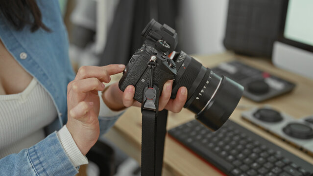 Woman adjusts camera dial with fingers while holding a mirrorless lens near keyboard and monitor in studio; focus creativity craft.