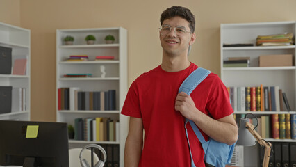 Man holding backpack strap with right hand, smiling in a building study room with bookshelves and desk, red shirt, glasses  confidence focus. © Krakenimages.com